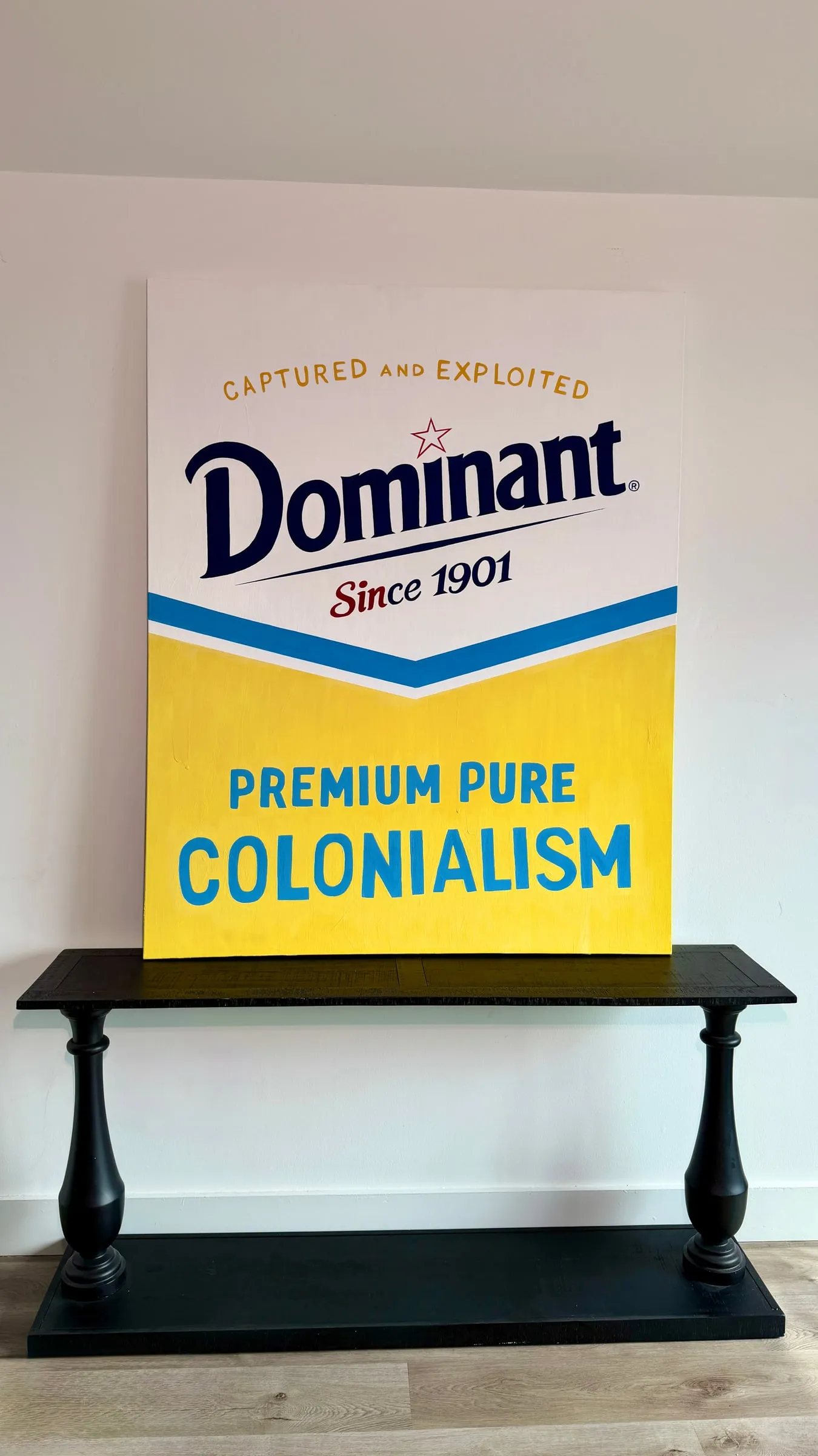 Painting of a yellow and white Dominant, Premium Pure Colonialism sign, standing on a black console table