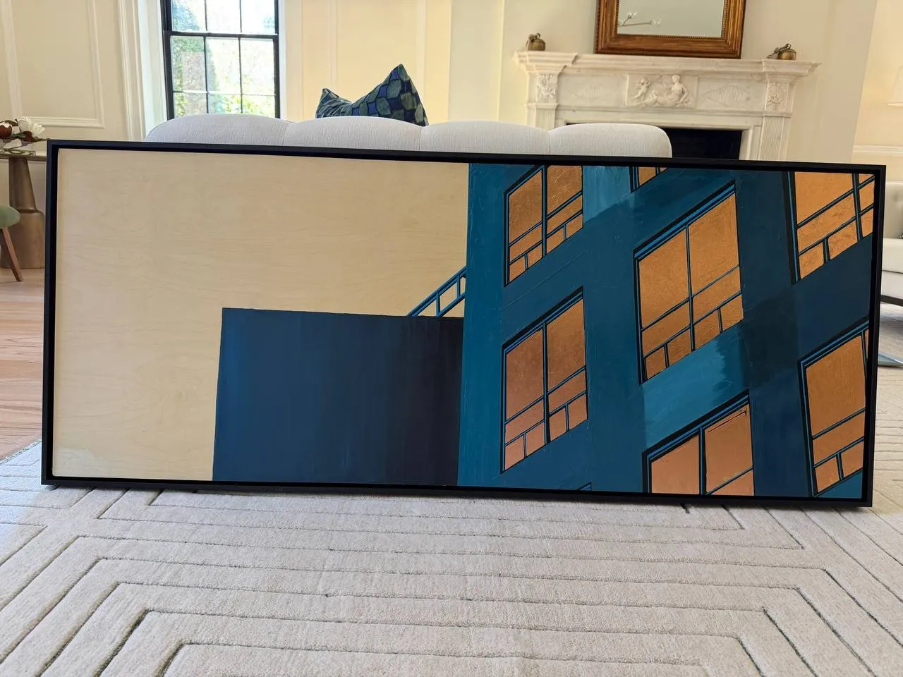 The framed print standing in a living room in front of a white sofa, with a marble fireplace behind