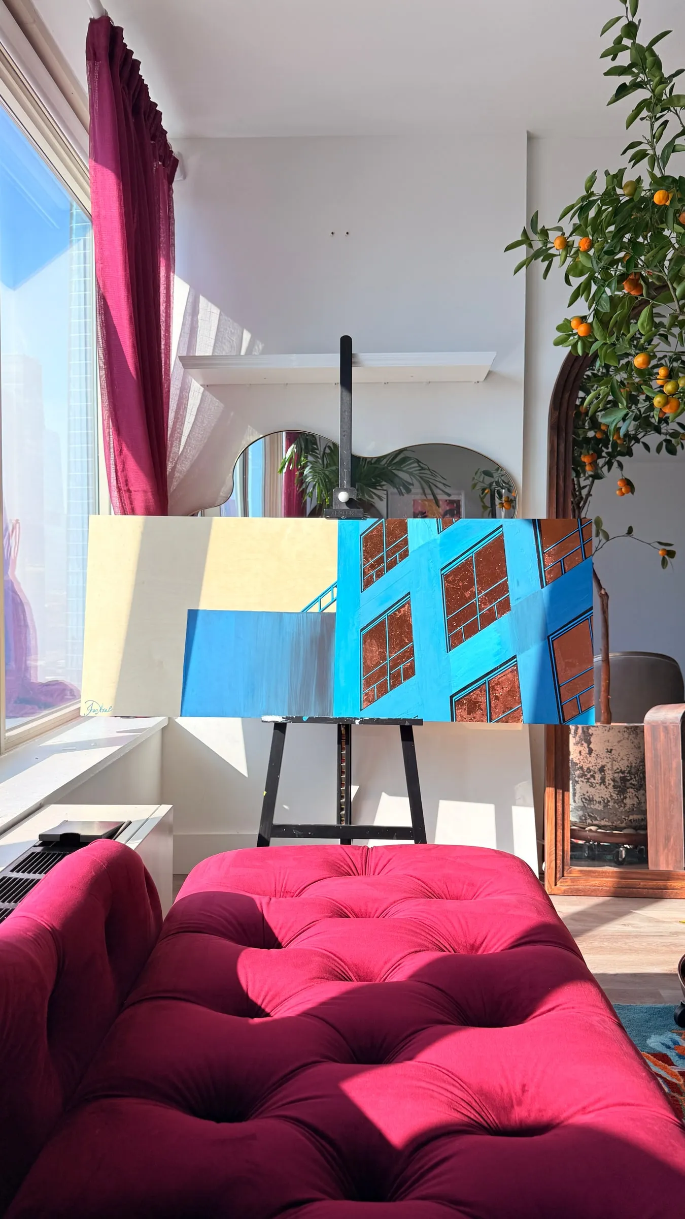 Blue and orange abstract canvas on an easel in a sunlit studio with a pink sofa