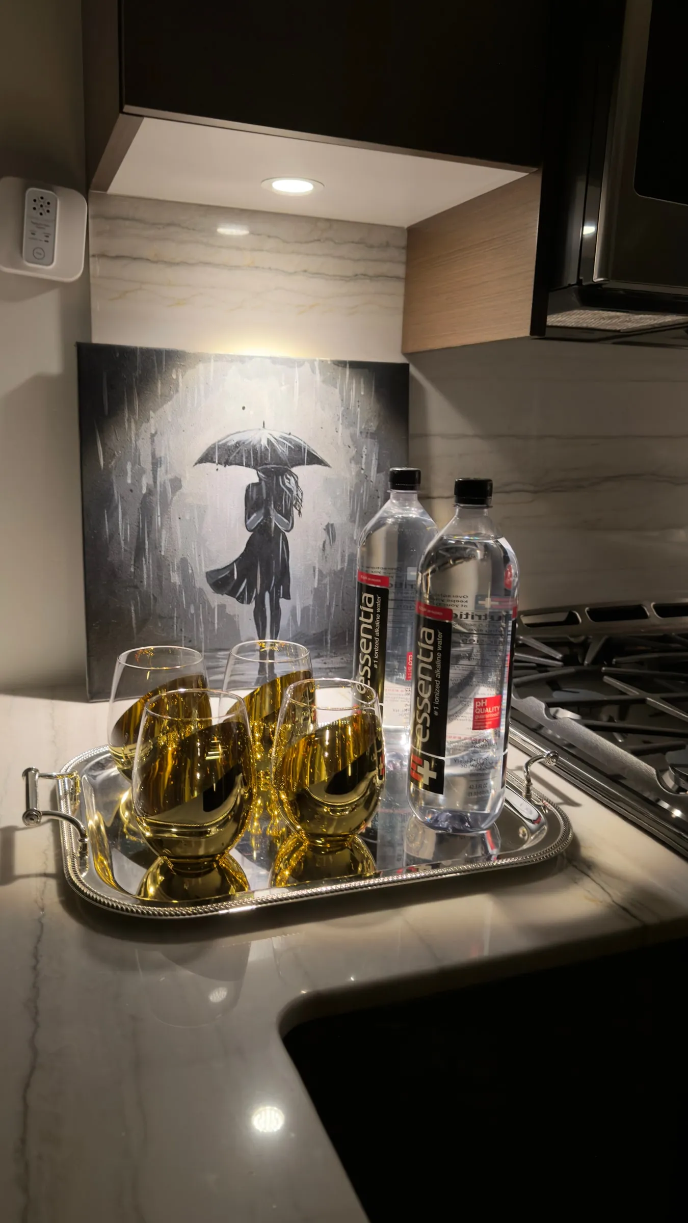 On a marble kitchen counter under a spotlight, behind a silver tray of gold glasses: monochrome painting of a woman under an umbrella in the rain