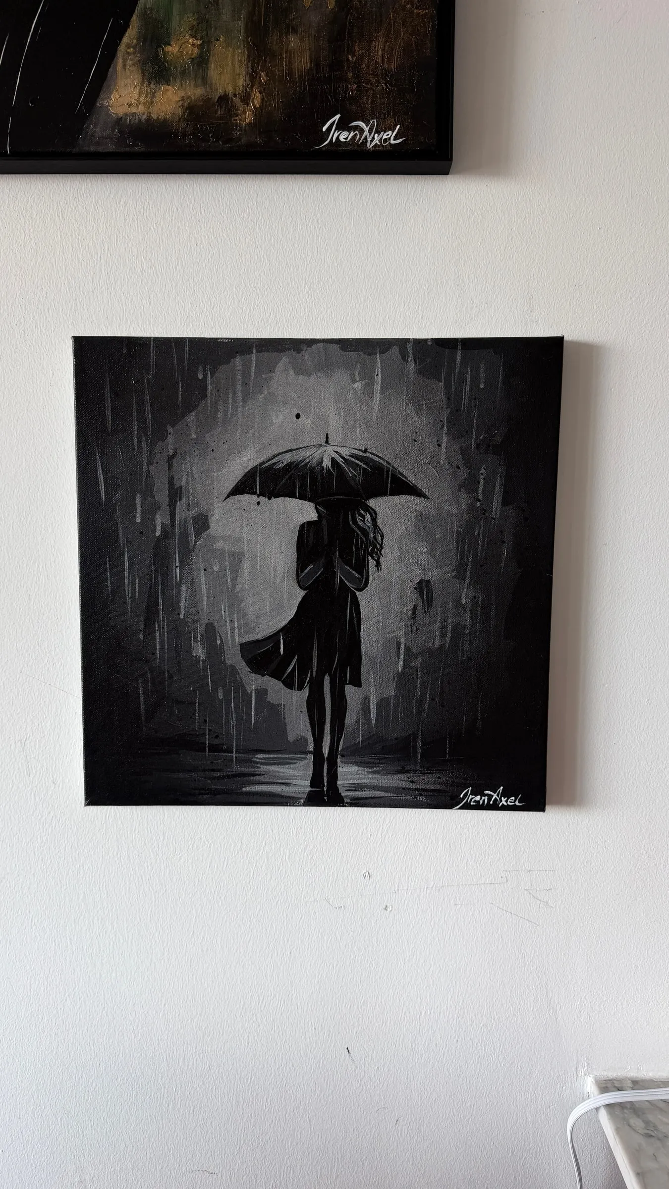 Hung on a white wall beneath a framed gold-and-black painting: monochrome painting of a woman under an umbrella in the rain